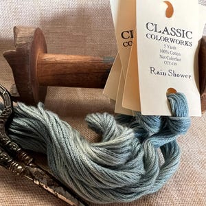 May include: A skein of blue-green cotton embroidery floss, labeled "Classic Colorworks Rain Shower" on a card, with a wooden spool and antique scissors.