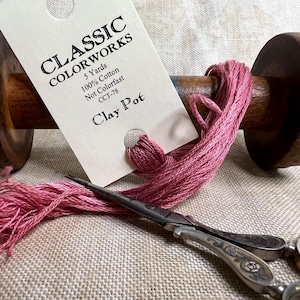 May include: A wooden thread spool with pink thread and a tag that reads "CLASSIC COLORWORKS Clay Pot." The spool is next to a pair of vintage scissors. The thread is 100% cotton and 5 yards.