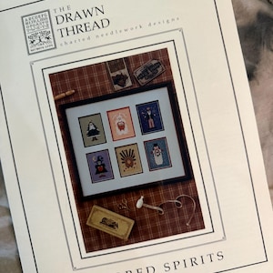 May include: A needlework pattern booklet titled The Drawn Thread Kindred Spirits featuring a framed collection of six seasonal cross-stitch designs including a Christmas tree, a turkey, and a snowman, displayed against a brown plaid background.