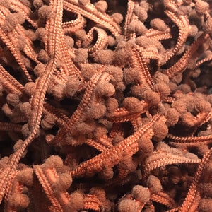 May include: A close-up of a pile of brown pom-pom trim. The trim is made of soft, fuzzy material and has a subtle sheen.