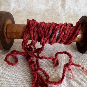 May include: A wooden spool with a red, fuzzy yarn wound around it. The yarn is loose and hanging off the spool.