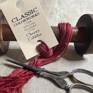 May include: A spool of red embroidery floss wound around a wooden bobbin. The floss is labeled "Classic Colorworks Cherry Cobbler CCT-227 5 Yards 100% Cotton Not Colorfast".