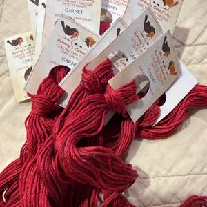 May include: A collection of deep red embroidery floss skeins, each attached to a white paper tag labeled Romy's Creations, Garnet, 6 stranded cotton, and 8.75 yd. The tags feature a small illustration of two birds and note the threads are not colorfast.