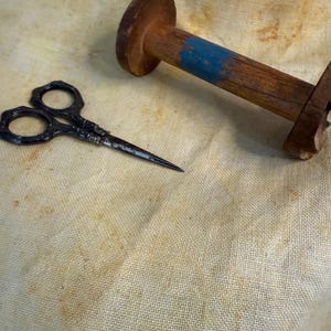 May include: An antique sewing kit with a pair of ornate scissors and a wooden spool. The scissors are black with decorative handles and a sharp point. The spool is brown with a blue stripe.