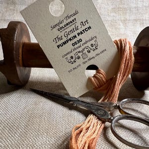 May include: A close-up of a brown wooden spool with a skein of peach-colored embroidery floss wrapped around it. A small card with the text "Sampler Threads COLORFAST by industry standards The Gentle Art PUMPKIN PATCH 0530 counted thread embroidery P.O. Box 670 New Albany, Ohio 43054" is attached to the spool. A pair of antique silver embroidery scissors are lying on a beige linen fabric.