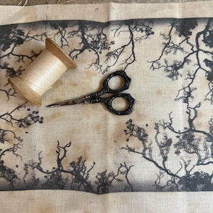 May include: A rectangular piece of beige fabric featuring a dark black printed border of bare tree branches. A wooden spool of light-colored thread and a pair of ornate black metal embroidery scissors are placed on top of the fabric.