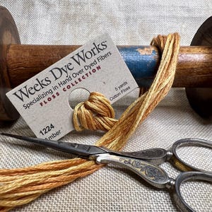 May include: A spool of amber colored cotton floss with a label that reads "Weeks Dye Works Floss Collection Specializing in Hand Over Dyed Fibers 1224 Amber 5 yards Cotton Floss".