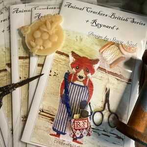 May include: A collection of cross-stitch patterns from the "Animal Crackers British Series" featuring a fox design. The image includes a wax embellishment, scissors, and spools of thread, suggesting a crafting theme.