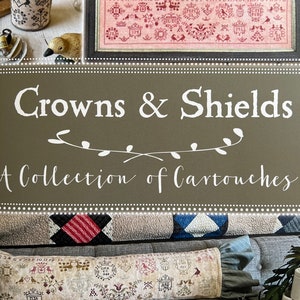 May include: A book cover with the title "Crowns & Shields: A Collection of Cartouches" by Blackbird Designs. The cover features a brown background with a white floral design and a pillow with a cross-stitch design.