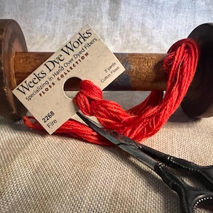 May include: A spool of red cotton floss labeled "Weeks Dye Works" with the color number "2268" and the name "Fire". The spool is wrapped in a brown wooden bobbin with metal ends. The floss is 5 yards long and is made of cotton.