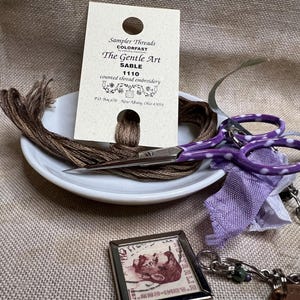 May include: A close-up of embroidery supplies including a spool of brown thread, a small white plate, a pair of purple scissors with white polka dots, a small purple fabric scrap, a silver chain, a small silver frame with a picture of a man, and a card with the text "Sampler Threads Colorfast The Gentle Art Sable 1110 counted thread embroidery P.O. Box 20 New Albany, Ohio 43054"