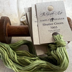 May include: Close-up of embroidery floss and tags. The floss is a vibrant green, and the tags read "Shutter Green" and "7003" with the brand name "The Gentle Art." A wooden spool and a jar of buttons are in the background.
