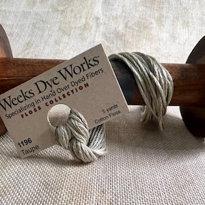May include: A spool of Weeks Dye Works floss in the color Taupe, 1196. The floss is a light brown color and is wound around a wooden spool. The label says "Weeks Dye Works" and "Specializing in Hand Over Dyed Fibers" and "FLOSS COLLECTION". The label also says "5 yards Cotton Floss".