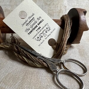 May include: A close-up of a brown and beige embroidery floss wound around a wooden spool. A card with the text "Sampler Threads COLORFAST The Gentle Art TORTOISE SHELL 1194 counted thread embroidery P.O. Box 670 New Albany, Ohio 43054" is visible. A pair of silver embroidery scissors are lying on top of the floss.
