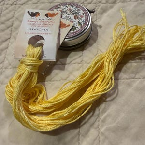 May include: A skein of bright yellow 6-stranded cotton embroidery thread labeled Sunflower by Romy's Creations, accompanied by a small round metal tin featuring a floral and bird pattern with the text Articles de Mercerie de Qualité.