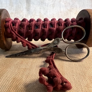 May include: A spool of burgundy pom-pom trim with a pair of antique silver scissors. The trim is wrapped around a wooden spool.