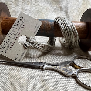 May include: A spool of beige cotton floss wound around a wooden bobbin. The spool is labeled "Weeks Dye Works Floss Collection" and "1173 Confederate Gray". A pair of antique silver scissors with floral designs are lying on a beige linen fabric.