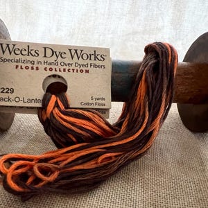 May include: A spool of Weeks Dye Works floss in the color "Jack-O-Lantern" with orange, brown, and black hand-dyed cotton threads. The label reads "Weeks Dye Works Specializing in Hand Over Dyed Fibers FLOSS COLLECTION 2229 Jack-O-Lantern 5 yards Cotton Floss".