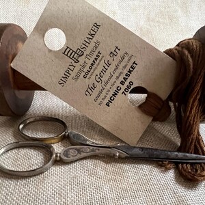 May include: A brown embroidery floss skein with a white tag that reads "SIMPLY SHAKER Sampler Threads COLORFAST The Gentle Art counted thread embroidery PO Box 670 New Albany, Ohio 43054 PICNIC BASKET 7060".