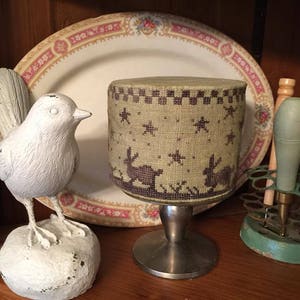 May include: A white ceramic bird figurine with a brown base, a beige and brown fabric covered cylinder with a star pattern, a white and gold plate with a floral design, and a green metal stand with three wooden dowels.