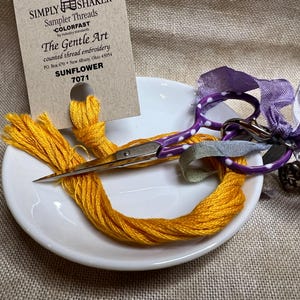 May include: A white ceramic dish with a spool of yellow embroidery floss, a pair of purple scissors with white polka dots, and a purple ribbon. The floss is labeled "Sunflower 7071" and "The Gentle Art counted thread embroidery".