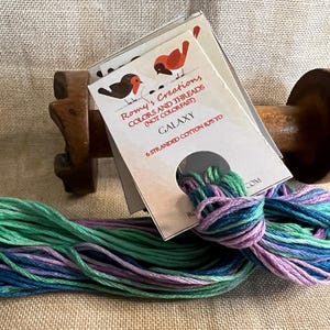 May include: A skein of six-strand cotton embroidery floss in shades of green, blue, and purple. The floss is wound around a wooden spool and labeled "Romy's Creations Colors and Threads (Not Colorfast) Galaxy 6 Stranded Cotton 875 yd."