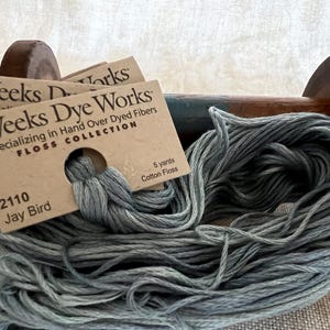 May include: A skein of gray cotton floss labeled "Weeks Dye Works" with the color name "Jay Bird" and the number "2110". The label also states "Specializing in Hand Over Dyed Fibers" and "Floss Collection".