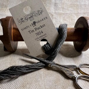 May include: A spool of gray embroidery floss wound around a wooden bobbin. A card with the text "Simply Shaker Tin Bucket 7024 Colorfast" is attached to the spool. A pair of silver scissors with ornate handles is also pictured.