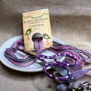 May include: A white ceramic dish with a pink and gray embroidery floss skein, purple polka dot scissors, and a purple ribbon. A card with the text "Romys Creations Colors and Threads Ocean Drive" is in the background.