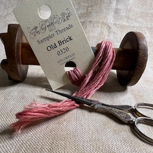 May include: A spool of pink embroidery floss wound around a wooden bobbin. The floss is labeled "The Gentle Art Sampler Threads Old Brick 0320 COLORFAST". A pair of silver embroidery scissors are also pictured.