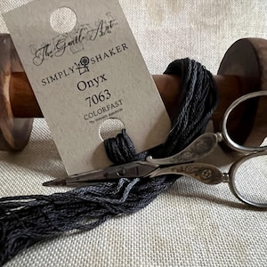 May include: A spool of black embroidery floss with a label that reads "The Gentle Art Simply Shaker Onyx 7063 Colorfast". The floss is wrapped around a wooden spool and a pair of antique scissors are nearby.
