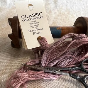 May include: A skein of mauve embroidery floss wound around a wooden spool with a label that reads "CLASSIC COLORWORKS 5 Yards 100% Cotton Not Colorfast CCT-427 Root Beer Float".