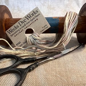 May include: A wooden yarn winder with a skein of multi-colored yarn wound around it. A pair of scissors and a card with the text "Weeks Dye Works" are also in the image. The card also says "Specializing in Hand Over Dyed Fibers" and "Holiday Collection".