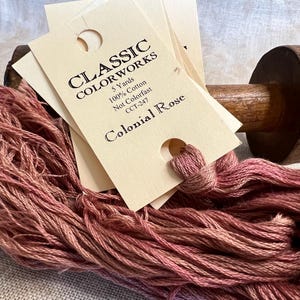 May include: A skein of Colonial Rose embroidery floss, a dusty rose color, is wrapped around a wooden spool. The floss is 100% cotton and is 5 yards long. The label reads "Classic Colorworks, 5 Yards, 100% Cotton, Not Colorfast, CCT-247, Colonial Rose."