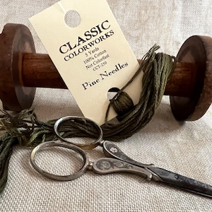 May include: A wooden spool of green thread with a label that reads "Classic Colorworks 5 Yards 100% Cotton Not Colorfast CCT-250 Pine Needles". A pair of antique silver embroidery scissors are also pictured.
