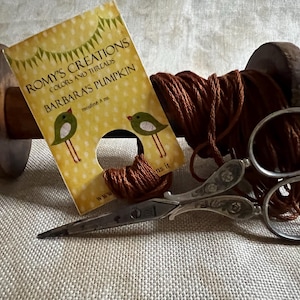 May include: A spool of brown thread with a label that reads "Romys Creations Colors and Threads Barbara's Pumpkin mercerie & co. www.romys.it" and a pair of silver scissors with floral designs.