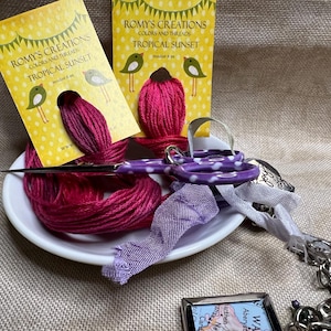 May include: Two packages of embroidery floss in the color "Tropical Sunset" by Romy's Creations. The floss is wound around cards with a yellow background and white polka dots. The cards feature a green bird illustration. The floss is on a white plate with a pair of purple scissors and a purple ribbon.