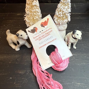 May include: A skein of Red Cherry embroidery floss with a label from Romy's Creations, featuring illustrations of birds. Two small ceramic dog figurines and two miniature Christmas trees are also in the image.
