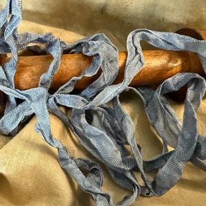 May include: A length of blue woven ribbon is draped over a wooden spool. The ribbon has a textured appearance and is arranged in loose loops. The spool is a light brown color and has a smooth finish. The background is a tan fabric.