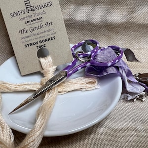 May include: A white ceramic dish with a skein of cream-colored embroidery floss and a pair of purple and white polka dot embroidery scissors. The dish is sitting on a beige fabric surface. A card with the text "SIMPLY SHAKER Sampler Threads COLORFAST The Gentle Art counted thread embroidery PO Box 670 New Albany Ohio 43054 STRAW BONNET 7002" is visible in the background.