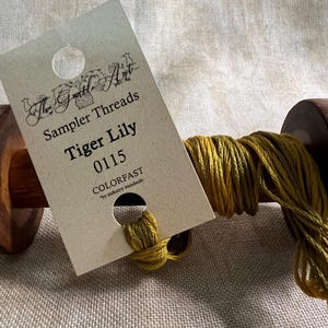 May include: A spool of green and yellow embroidery floss with a label that reads "The Gentle Art Sampler Threads Tiger Lily 0115 COLORFAST *by industry standards*"