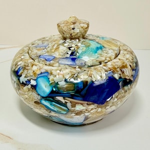 May include: A round, lidded container featuring a mosaic surface composed of embedded crushed white shells and larger, polished blue and turquoise stone fragments. The lid includes a small, central knob handle, and the entire exterior is finished with a smooth, glossy, clear resin coating.