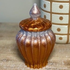May include: A decorative jar with a lid, featuring a metallic gold and silver glitter finish. The jar has a ribbed design and a pointed finial on the lid. The background includes a patterned ceramic container.