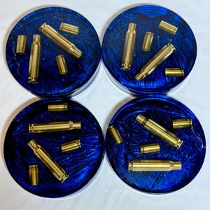 May include: Four round coasters with a deep blue, swirled resin design. Each coaster features brass bullet casings embedded within the resin. The coasters are circular and have a clear edge.
