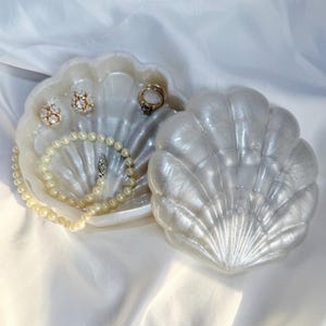 May include: A pearlescent white shell-shaped trinket dish with a matching lid. The open dish contains a string of cream-colored pearls, two round stud earrings with clear stones, and a gold ring with a dark stone set in the center of the dish.