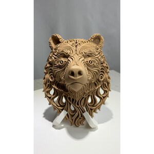 May include: A detailed, light brown bear head sculpture. The bear's fur is intricately designed with swirling patterns. The sculpture is mounted on a white stand, showcasing the bear's face and upper chest. The artwork is a decorative piece.