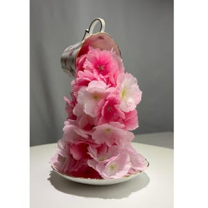 May include: A white porcelain teacup and saucer set featuring a delicate floral pattern. The teacup is tilted, appearing to pour a cascading arrangement of vibrant pink and light pink artificial cherry blossom flowers onto the saucer below.