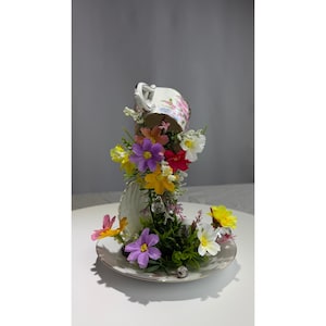 May include: A decorative teacup and saucer arrangement. The teacup is inverted, supported by the saucer, and filled with colorful artificial flowers. The flowers include yellow, purple, red, and white blooms, creating a whimsical display.