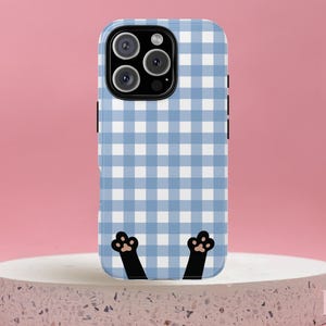 May include: A blue and white gingham patterned phone case with black cat paws at the bottom. The phone case is on a white stone pedestal against a pink background.