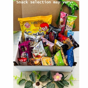 May include: A white box filled with a variety of individually wrapped snacks, including chips, chocolate wafers, and candies. The box is decorated with artificial flowers and greenery. Text at the top reads "Snack selection may vary."